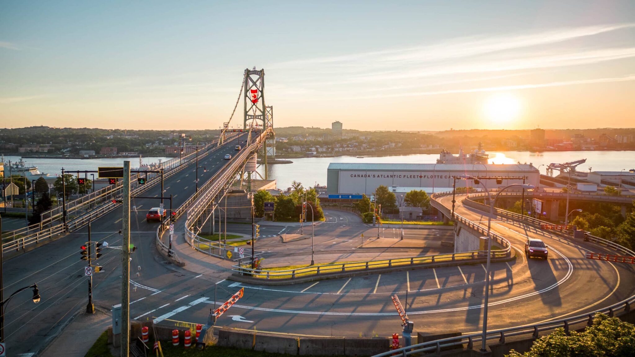 A vehicle drives on the Barrington Street ramp to the Macdonald Bridge at sunset. The Canadian flag flies in the tower of the bridge in the background.