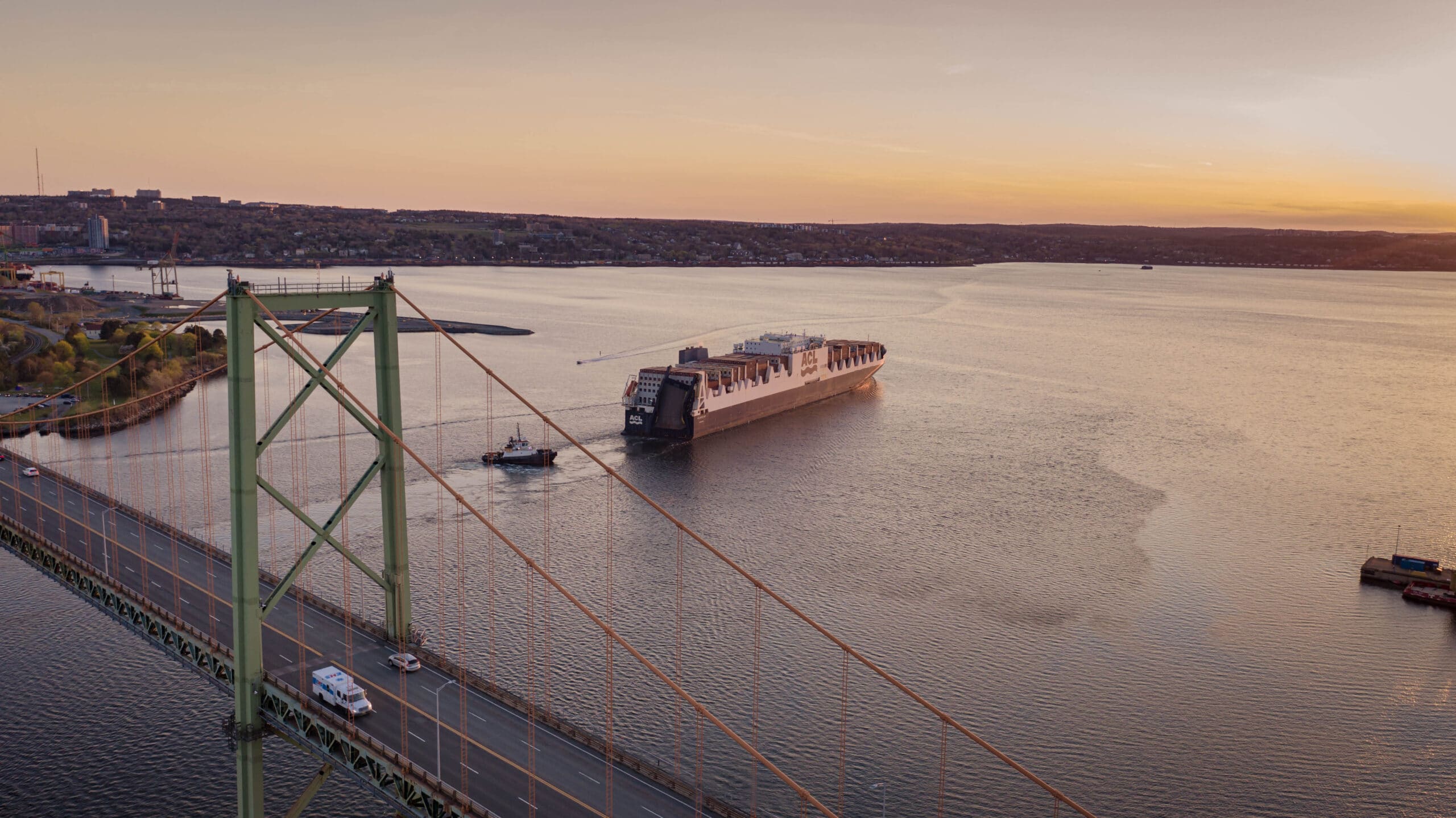 A container ship, accompanied by a tug boat, enters the Bedford Basin under the MacKay Bridge at sunset.