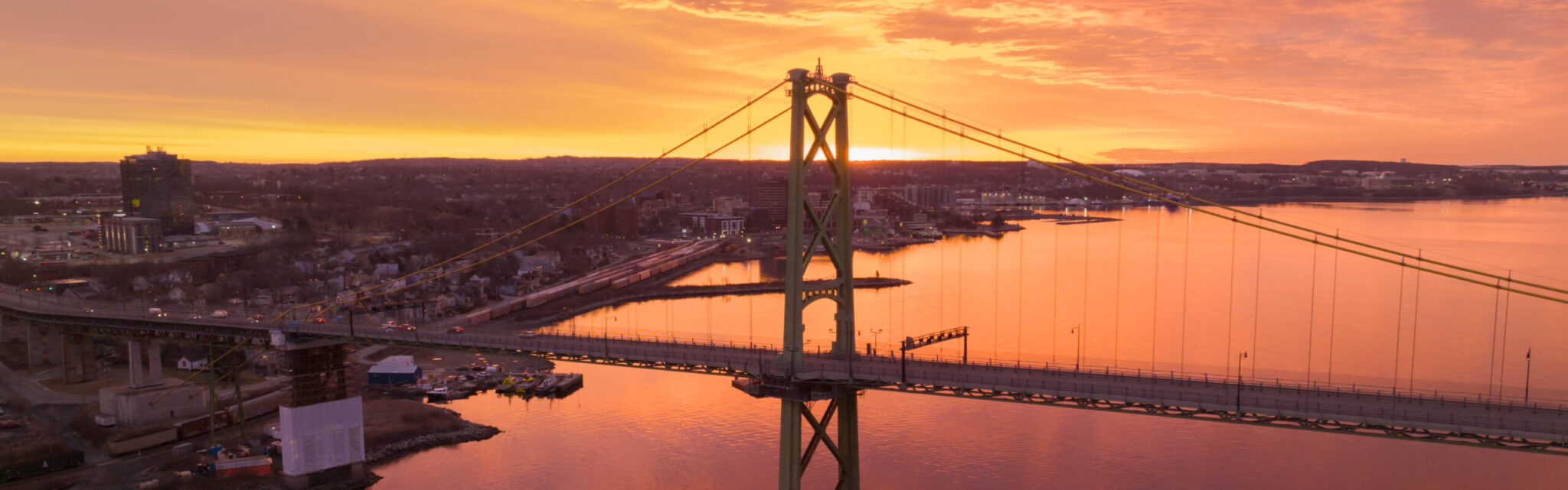 The Macdonald Bridge and Halifax Harbour are seen amid a red-orange sky at sunset.