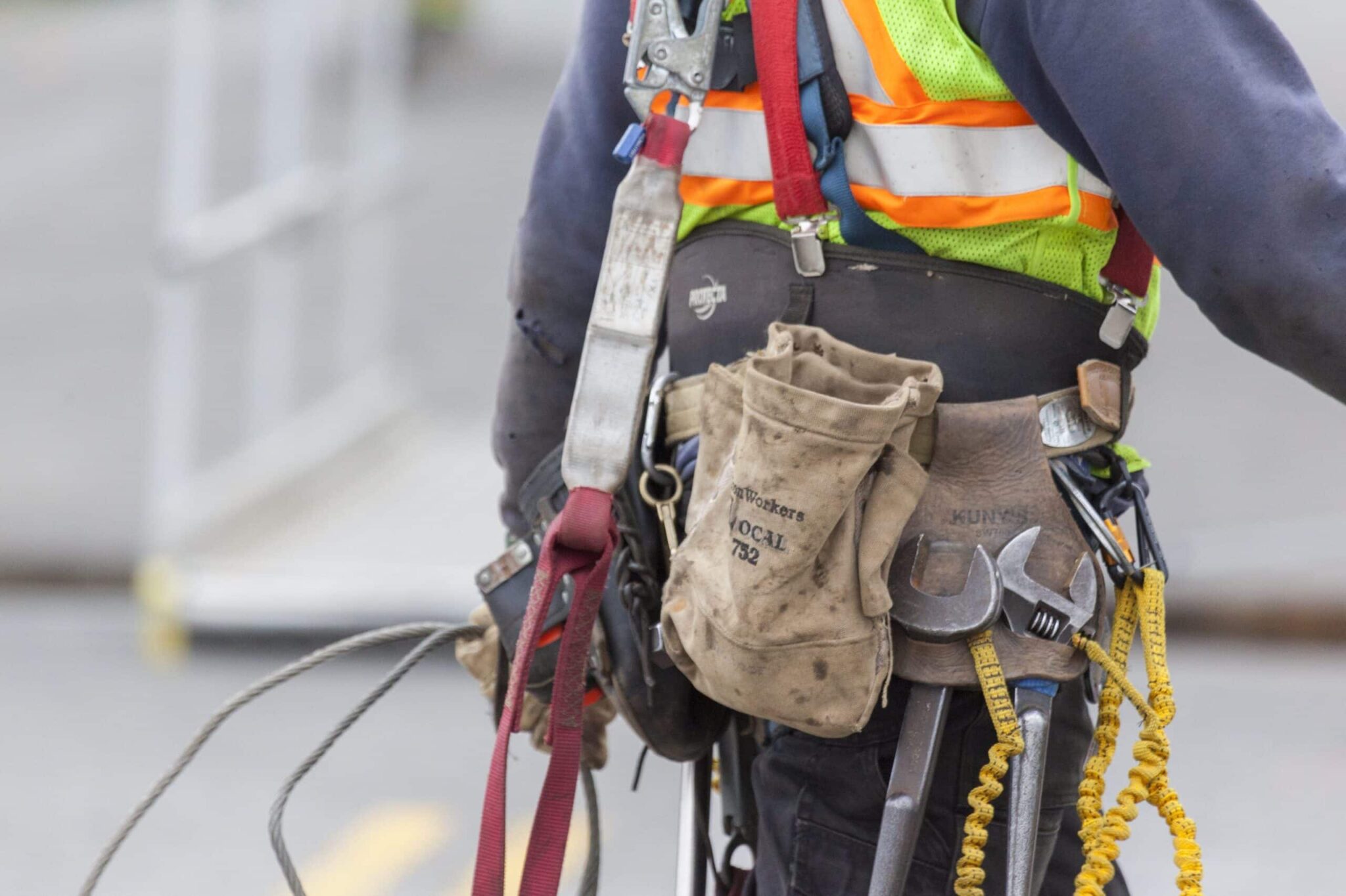 Tools and a bag are seen hanging from construction worker's tool belt. The worker is wearing a reflective safety vest.