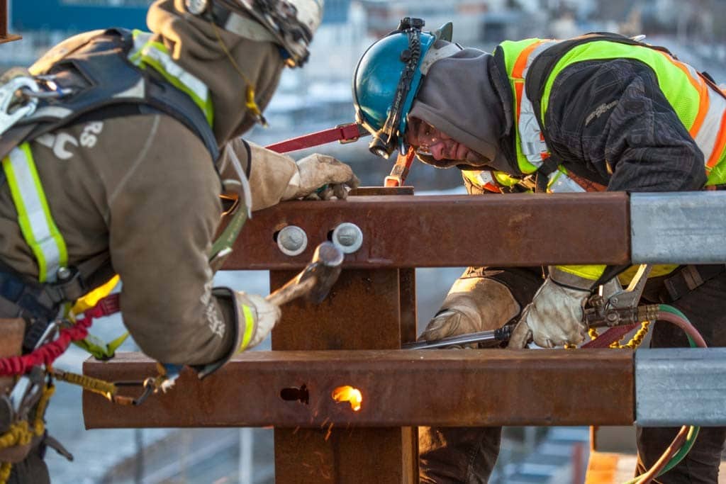 Construction workers work on steel components of the Macdonald Bridge in winter. The workers are bundled up and wearing reflective vests and hard hats.